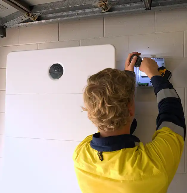 Technician in hi-vis workwear wiring a control box on a garage wall beside a home battery