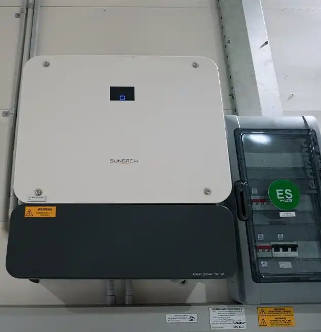 Sungrow inverter mounted on a wall beside a labelled switchboard with shutdown procedure and warning signs